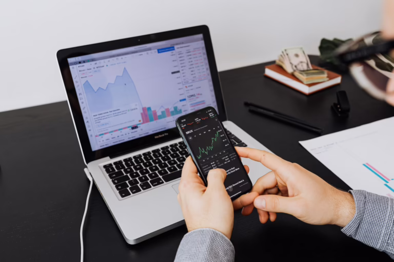 A person analyzes financial data using a smartphone and laptop, both displaying stock charts for SBUX (Starbucks Corporation), with visible line graphs, bar charts, and performance indicators. A stack of U.S. dollar bills, a pen, and printed financial sheets rest on the desk, symbolizing Algorithmic Stock Trading Automation and Market Insights through real-time data feeds, mobile-first analysis, and founder-led trading systems that empower investors to automate strategies, interpret market signals, and scale portfolio performance with precision.