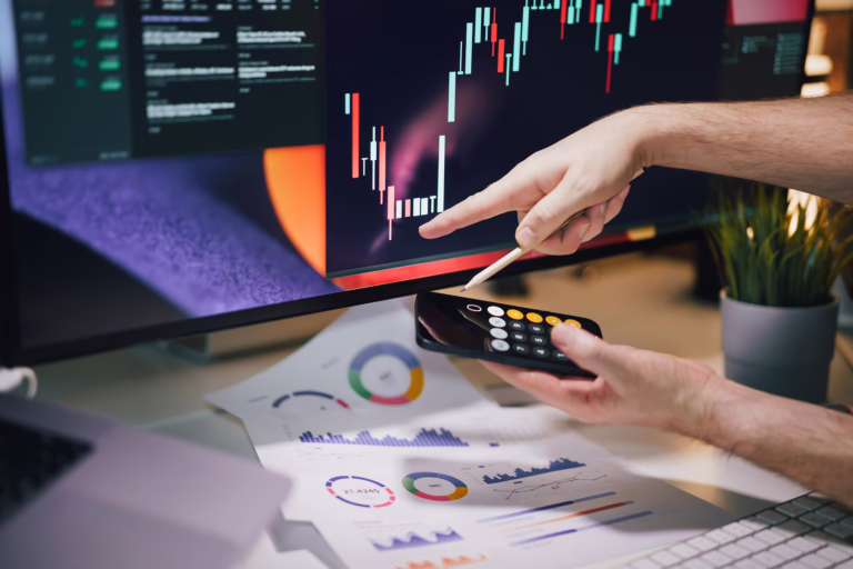 A person analyzes a candlestick chart on a computer monitor while pointing with a pencil and holding a smartphone displaying a calculator app. The desk is covered with printed financial charts, including pie, bar, and line graphs, alongside a keyboard and potted plant, symbolizing Bitcoin Price Tracking Tools for Smarter Decisions through founder-led crypto analytics, real-time data visualization, and strategic decision-making frameworks that empower investors to interpret market signals, optimize timing, and scale digital asset performance with precision.