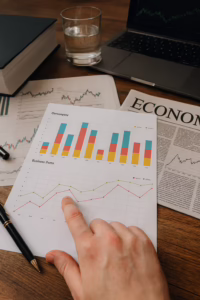 A workspace displays financial documents with colorful bar charts titled “Our company” and a line graph titled “Business Items,” showing monthly data trends for categories like Default, GTMU, and Students. A hand points to a specific data point on the line graph, while surrounding items include a newspaper with the headline “ECONOMY,” a laptop showing a stock market chart, a thick book, a pen, and a glass of water, symbolizing Economic Content Strategy for Authority and Growth through founder-led data analysis, editorial planning, and financial storytelling that empower businesses to build topical authority, interpret market signals, and drive sustainable growth.
