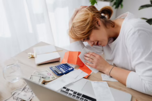 A person sits at a desk cluttered with receipts, a blue calculator, U.S. dollar bills, a notebook, and a laptop, holding financial documents with one hand while resting their head on the other, symbolizing Mastering Economic Stress Management through founder-led budgeting strategies, emotional resilience frameworks, and practical financial tools that empower individuals to navigate uncertainty, reduce money-related anxiety, and build sustainable habits for long-term economic stability.