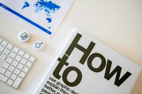 A creative workspace featuring a book titled “How to” alongside a world map, graphic design pins, and a keyboard, symbolizing Stock Market Tips for Small Businesses: Strategies for Stability and Growth through visual storytelling, global market awareness, and design-driven approaches to financial education and strategic planning.